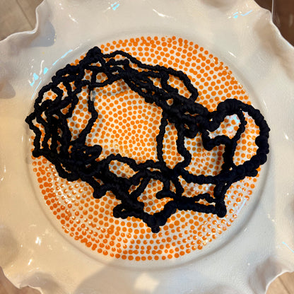 A black knitted or crocheted scarf with a wavy, organic pattern, laid flat on top of a circular white plate decorated with an orange dotted pattern.
