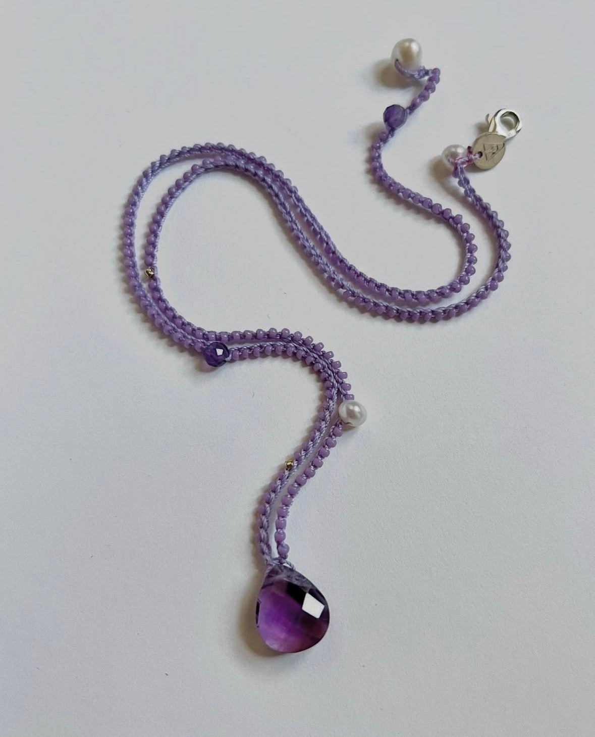 A purple beaded necklace with a faceted purple teardrop pendant, white pearl beads, and a silver lobster clasp, displayed on a plain light gray background.