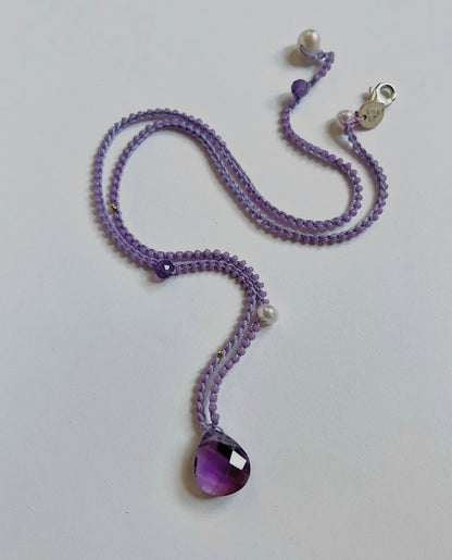 A purple beaded necklace with a faceted purple teardrop pendant, white pearl beads, and a silver lobster clasp, displayed on a plain light gray background.