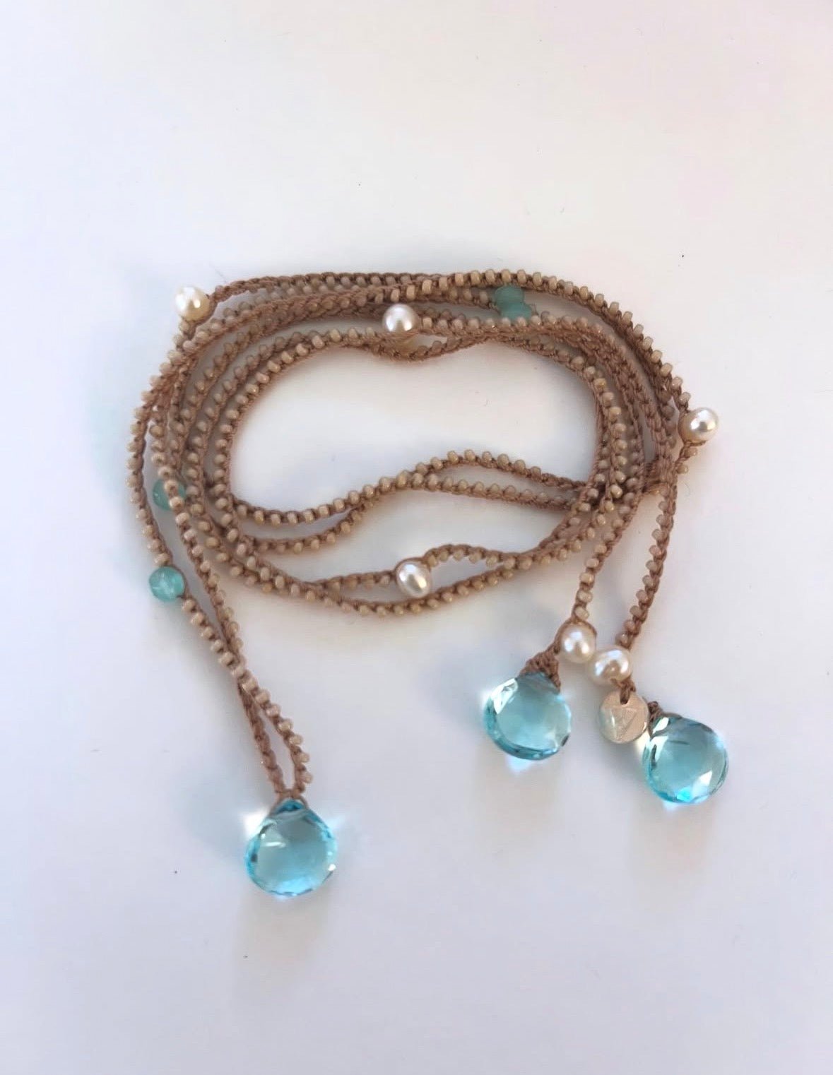 A multi-strand beaded necklace with brown woven cords, small white pearl-like beads, and light blue faceted teardrop pendant drops on a plain white background.