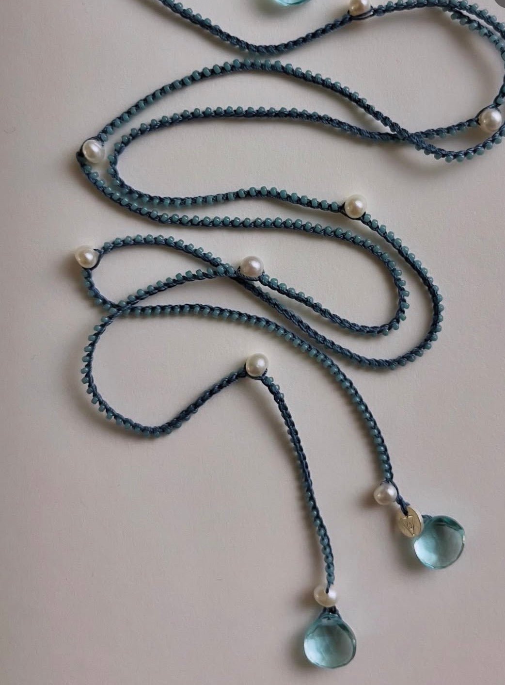A long braided cord necklace in a muted blue-gray color, decorated with small white pearl beads and light blue teardrop glass pendants, laid out on a plain light beige surface.