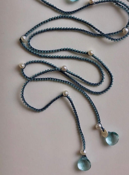 A long braided cord necklace in a muted blue-gray color, decorated with small white pearl beads and light blue teardrop glass pendants, laid out on a plain light beige surface.