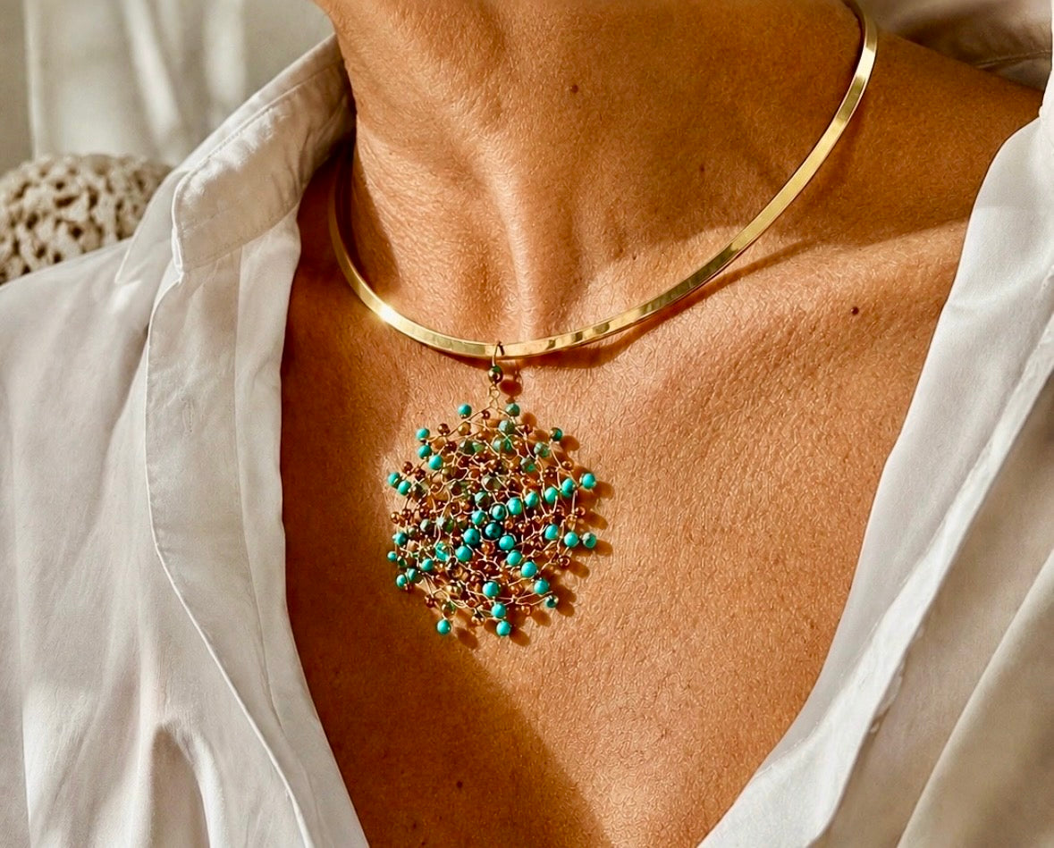 Gold-plated stainless steel choker necklace with a handwoven 18kt gold-plated copper wire Mandàla pendant featuring turquoise and bronze crystal beads, worn over a white linen shirt.