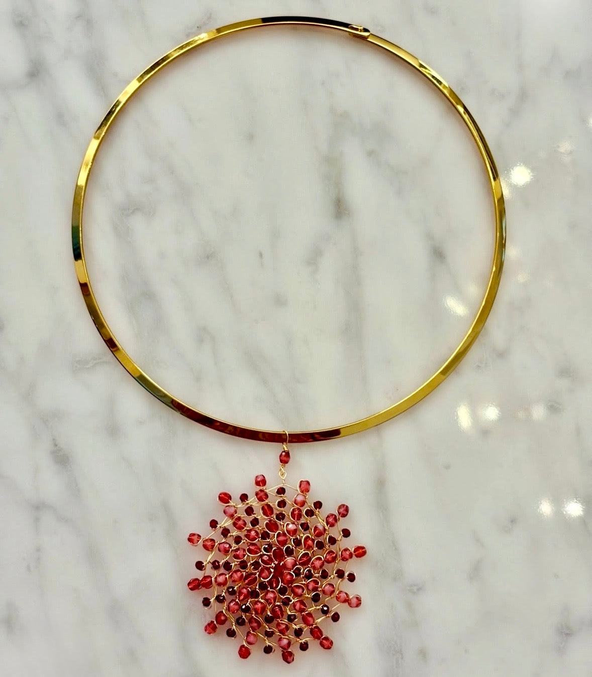 Gold-plated stainless steel rigid choker necklace with a handwoven 18kt gold-plated copper wire Mandàla pendant featuring ruby and garnet crystal beads, displayed on white marble.