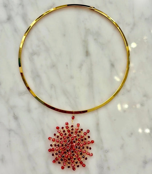 Gold-plated stainless steel rigid choker necklace with a handwoven 18kt gold-plated copper wire Mandàla pendant featuring ruby and garnet crystal beads, displayed on white marble.
