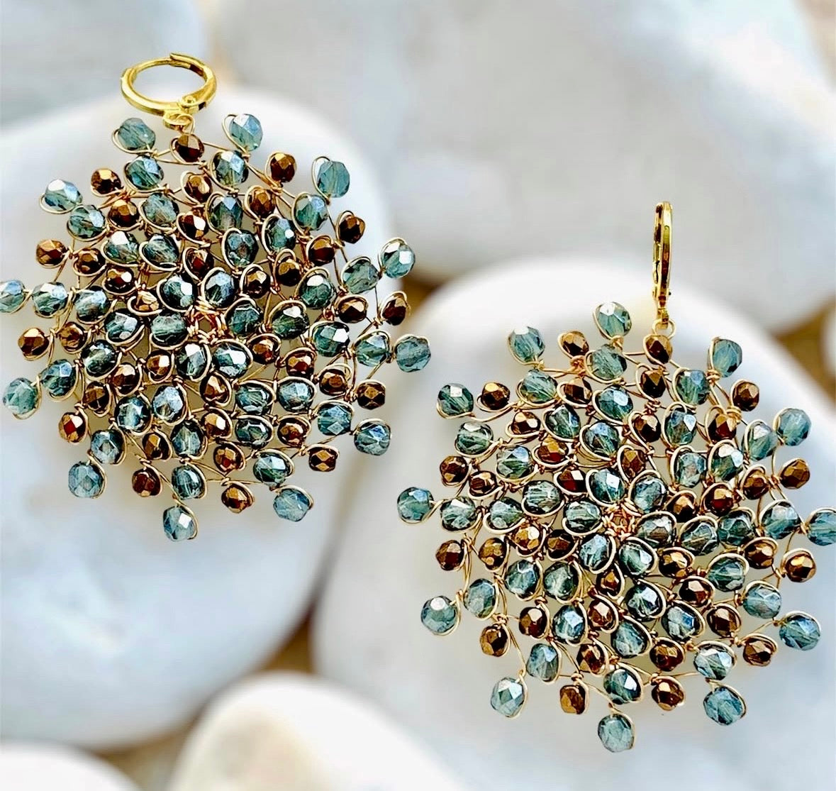 Handwoven gold-plated copper wire Mandàla earrings with sky blue and bronze Bohemian crystals, displayed on white pebbles.