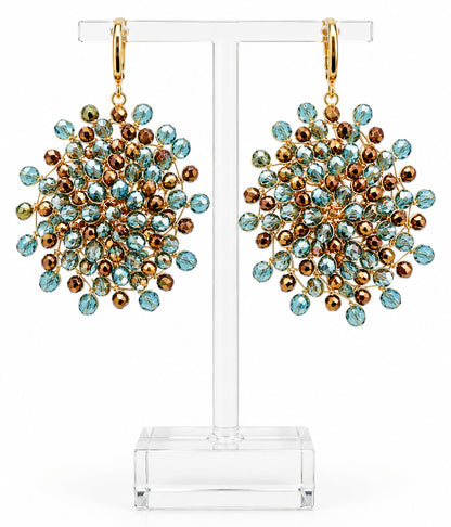 Boucles d'oreilles Mandàla tissées main en fil de cuivre plaqué or avec cristaux de Bohême bleu ciel et bronze, posées sur des galets blancs.