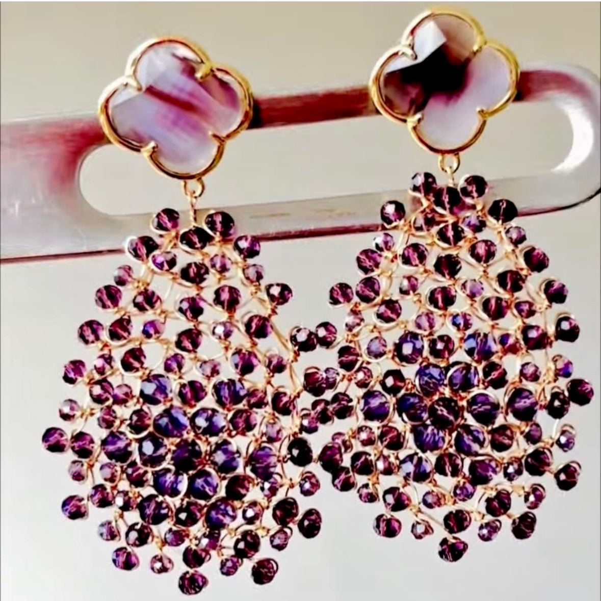 Handwoven 18kt gold-plated copper wire Mandàla earrings with faceted violet Bohemian crystals and a clover-shaped crystal stud top, displayed on a jewellery stand.