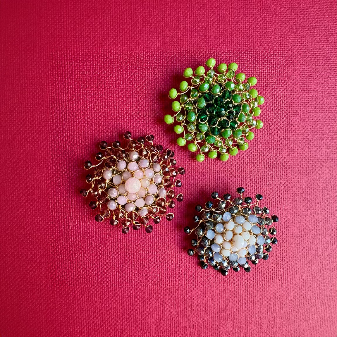 Three handmade gold wire floral brooches with faceted beads, arranged on a textured hot pink background. One brooch features light pink beads, one features dark green beads, and the third features light blue and white beads.