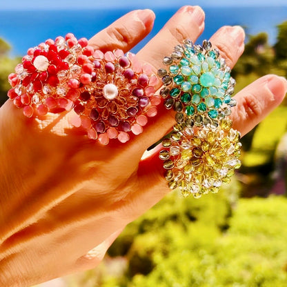 Four handwoven Mandàla rings in red, pink, teal and olive green with champagne crystals, displayed on a hand against a sunny outdoor background.