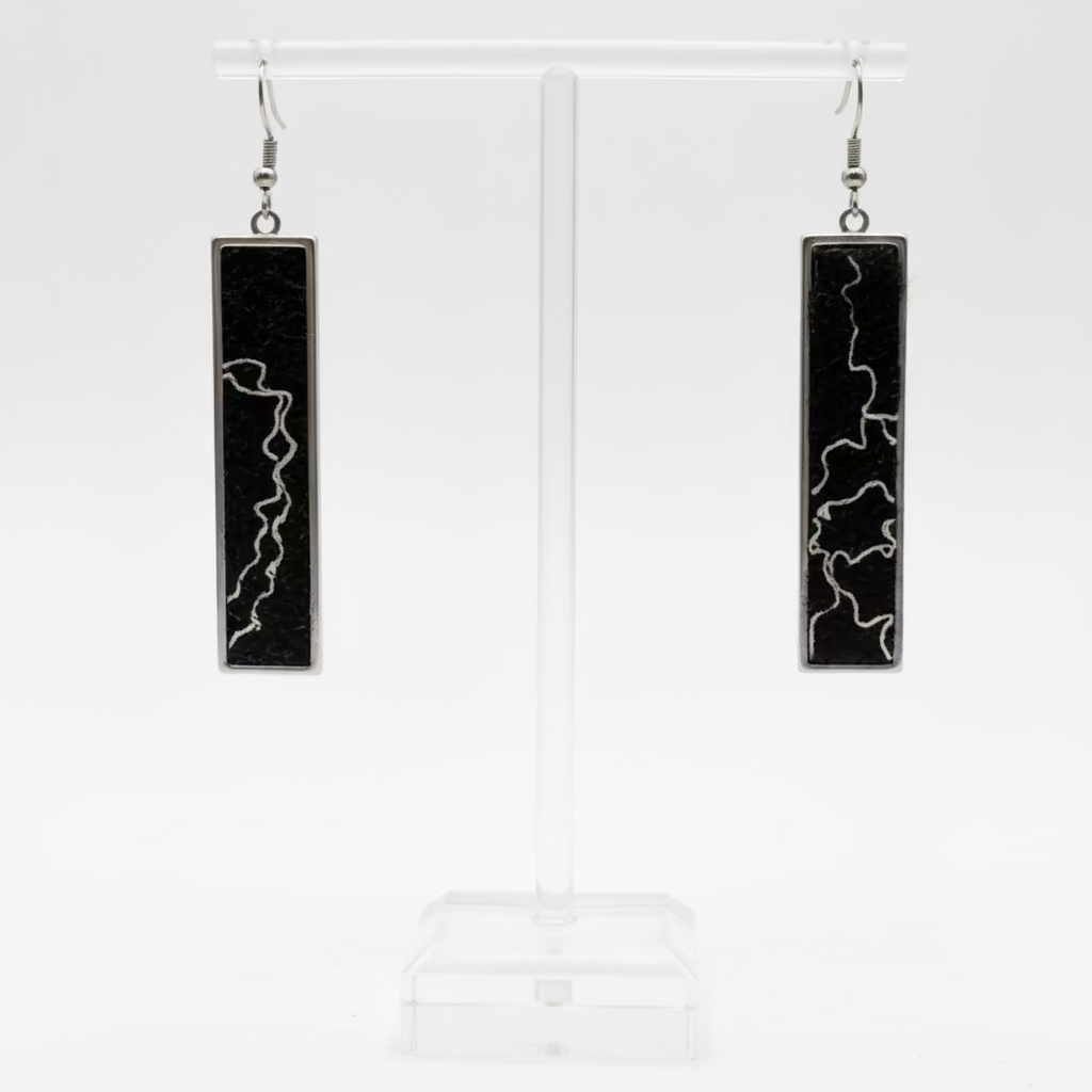 A pair of long rectangular pendant earrings with black inlay and white lightning-bolt patterns, displayed on a clear acrylic stand against a white background.
