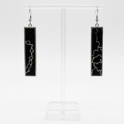 A pair of long rectangular pendant earrings with black inlay and white lightning-bolt patterns, displayed on a clear acrylic stand against a white background.