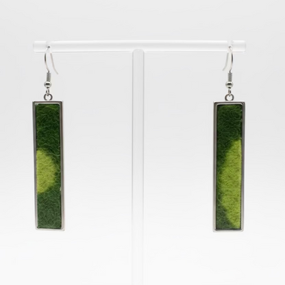 A pair of long rectangular pendant earrings with olive green and lime inlay and silver-toned metal borders, displayed on a clear acrylic stand against a white background.
