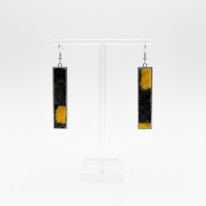 A pair of long rectangular drop earrings with a black and yellow abstract pattern from Nuno Felt, displayed on a clear acrylic stand against a white background.