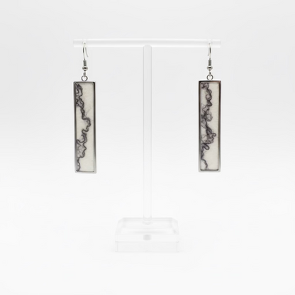 A pair of rectangular long pendant earrings with dark abstract marbled patterns on a cream background, displayed on a clear acrylic stand with a white background.