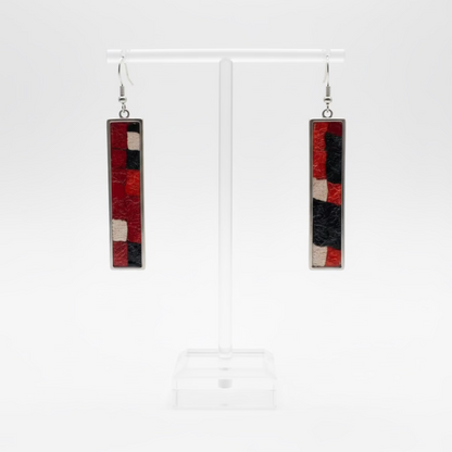 A pair of long rectangular dangle earrings with a red, black, and cream geometric pattern from Nuno Felt, displayed on a clear acrylic stand against a white wall.