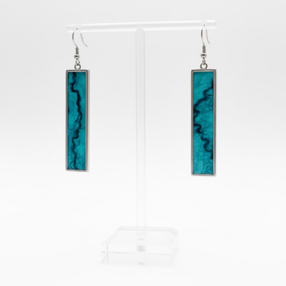 A pair of rectangular drop earrings with turquoise blue and black marbled inlay and silver-toned metal borders, displayed on a clear acrylic stand against a white textured wall.