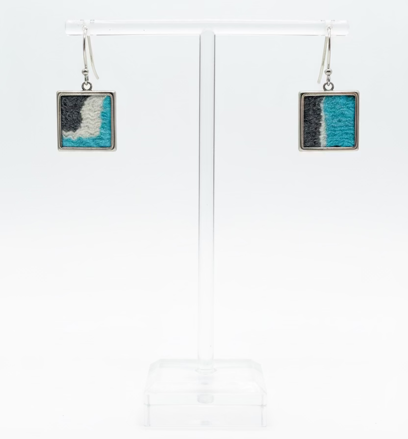 A pair of square drop earrings displayed on a clear acrylic stand. Each earring has a silver-toned metal frame with a textured inlay of turquoise blue, black, and white.