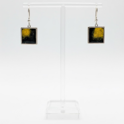 A pair of square drop earrings with a textured yellow and black Nuno Felt center and silver-tone borders, displayed on a clear acrylic stand against a white background.