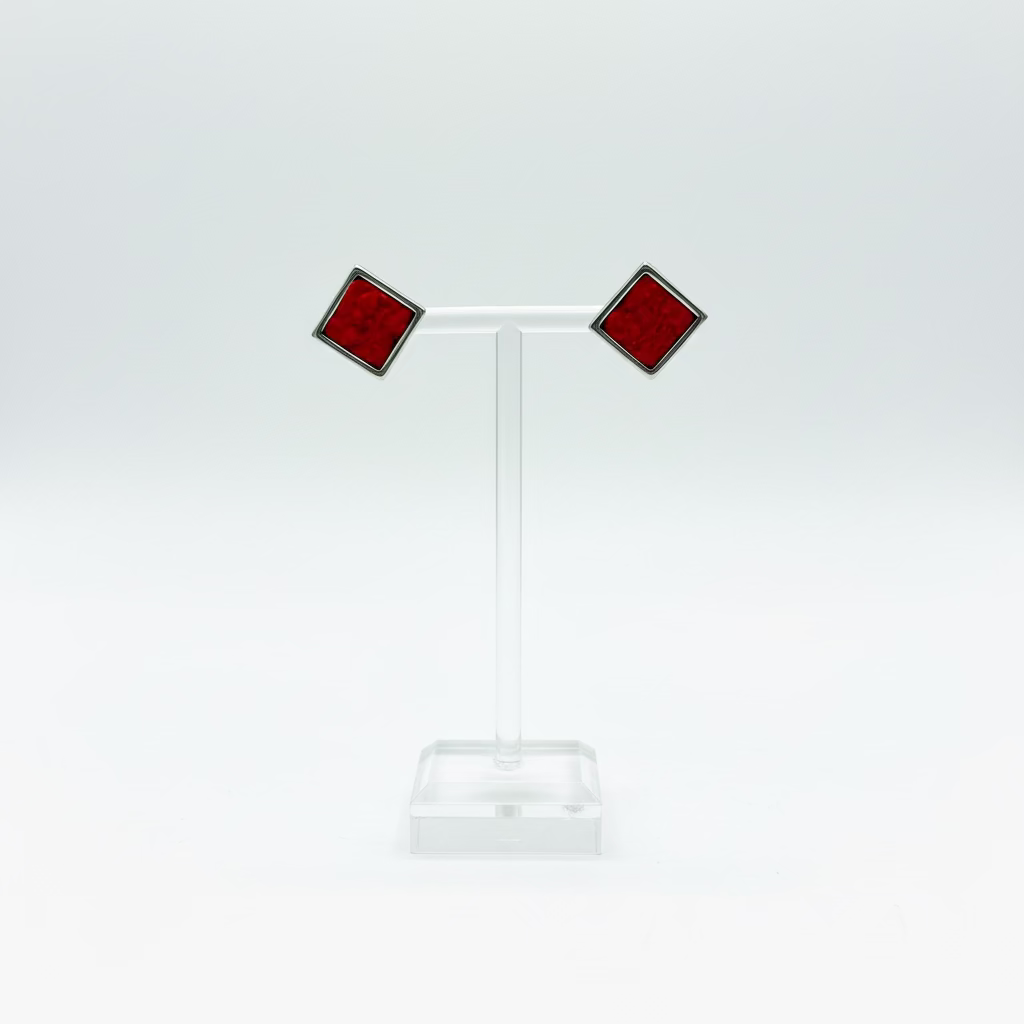 A pair of red square stud earrings with dark metal borders displayed on a clear acrylic T-stand against a white background.