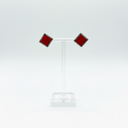 A pair of red square stud earrings with dark metal borders displayed on a clear acrylic T-stand against a white background.