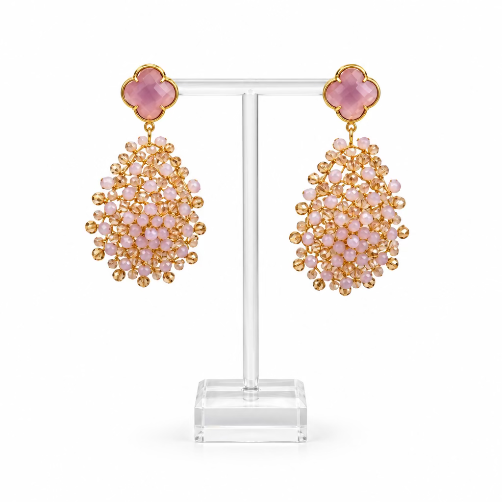 Une paire de boucles d'oreilles pendantes statement sur un présentoir en acrylique transparent. Chacune présente un haut rose en forme de trèfle bordé de doré et, en dessous, une grande grappe de petites perles facettées rose pâle, le tout en métal doré.
