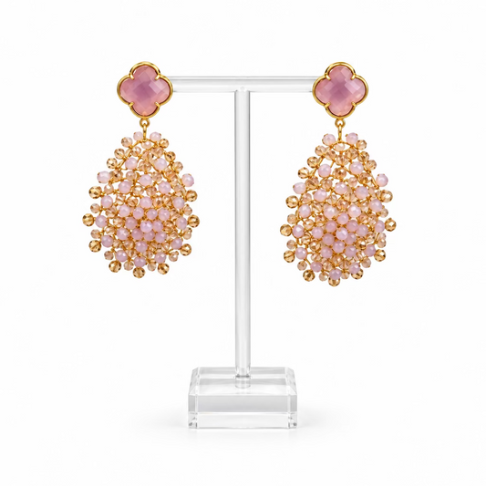 A pair of dangling statement earrings displayed on a clear acrylic stand. Each earring has a pink clover-shaped top with a gold-tone border and a large cluster of small pale pink faceted beads below, with gold-tone metal throughout.