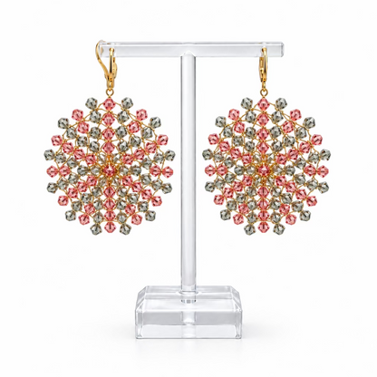 Une paire de boucles d'oreilles Mandala en cristaux Swarovski rose et gris-vert enfilés sur fil plaqué or, sur fond blanc.
