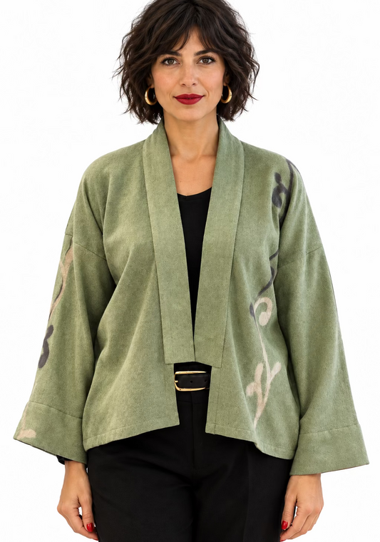 A dark haired woman wearing a sage coloured kimono merino wool lightweight jacket with a black top and black trousers.  The jacket has a fleur d'alis grey and cream pattern down the sleeve.