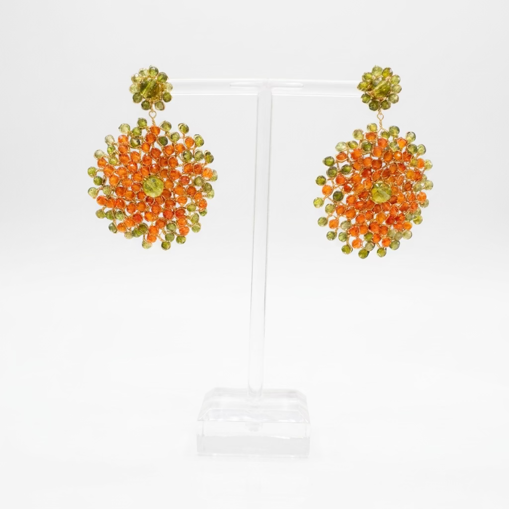 A pair of handmade  dangle earrings displayed on a clear acrylic stand. The earrings feature a dense cluster of orange and light green crystals with a small green crystal at the center.