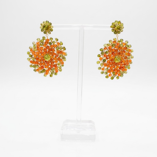 A pair of handmade  dangle earrings displayed on a clear acrylic stand. The earrings feature a dense cluster of orange and light green crystals with a small green crystal at the center.