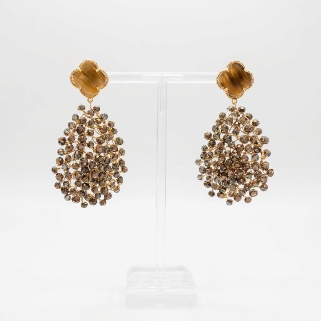 A pair of statement dangle earrings displayed on a clear acrylic stand. The earrings feature gold-toned clover leaf studs with large, teardrop-shaped clusters of bronze crystals.