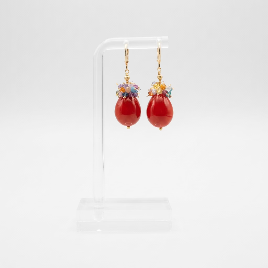 A pair of red lacquered Maiorca pearl dangle earrings with colourful bunched tourmalines with  gold hooks, displayed on a clear acrylic stand in a softly blurred indoor setting.