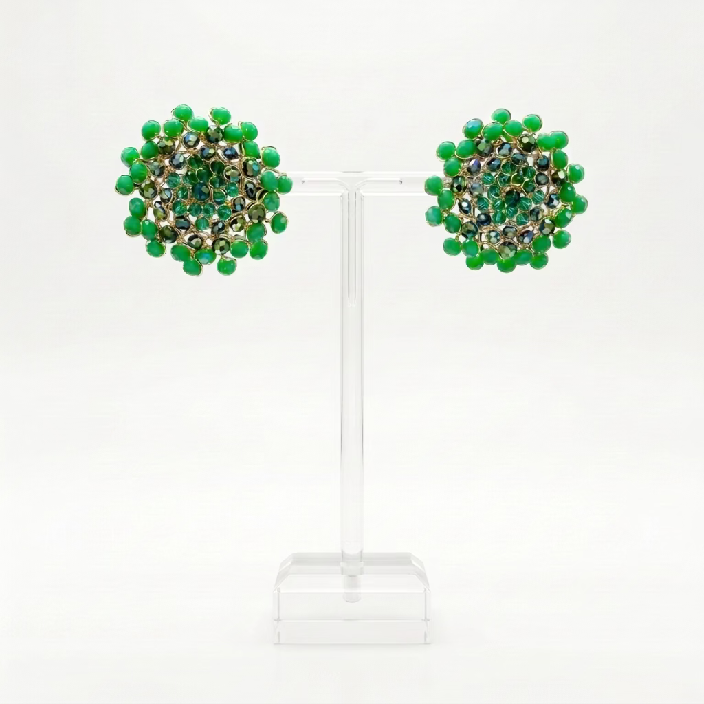 A round pair of button mandala stud earrings made with a burst of faceted green crystals in three layers. The outer ring is bright, opaque leaf green, the middle ring flashes deep forest green with an iridescent sheen, photographed on a perspex stand on a white background.