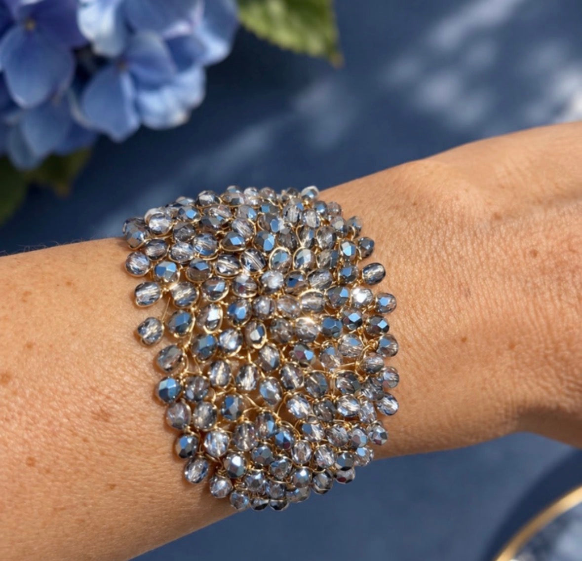 A wide cuff bracelet covered in faceted Bohemian lavender crystals set in gold-toned metal, worn on a person's wrist against a dark blue background with blurred blue flowers in the corner.