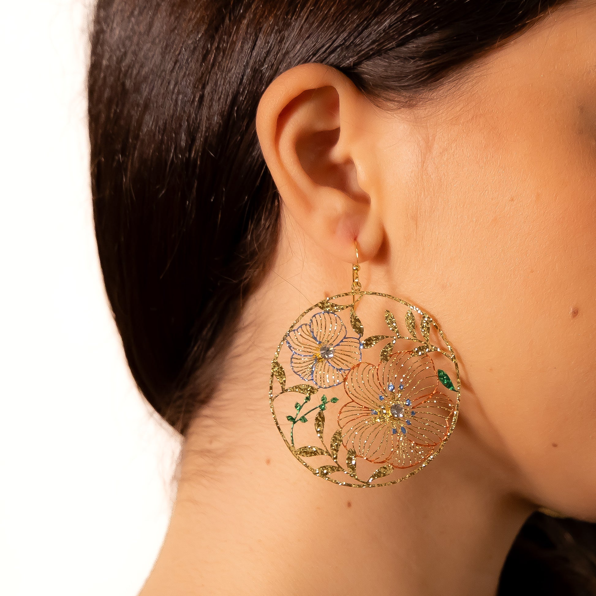 A woman wearing large round brass drop earrings with openwork floral design, hand-painted enamel in blue, coral and green, crystal centres and gold hook fastening on white background