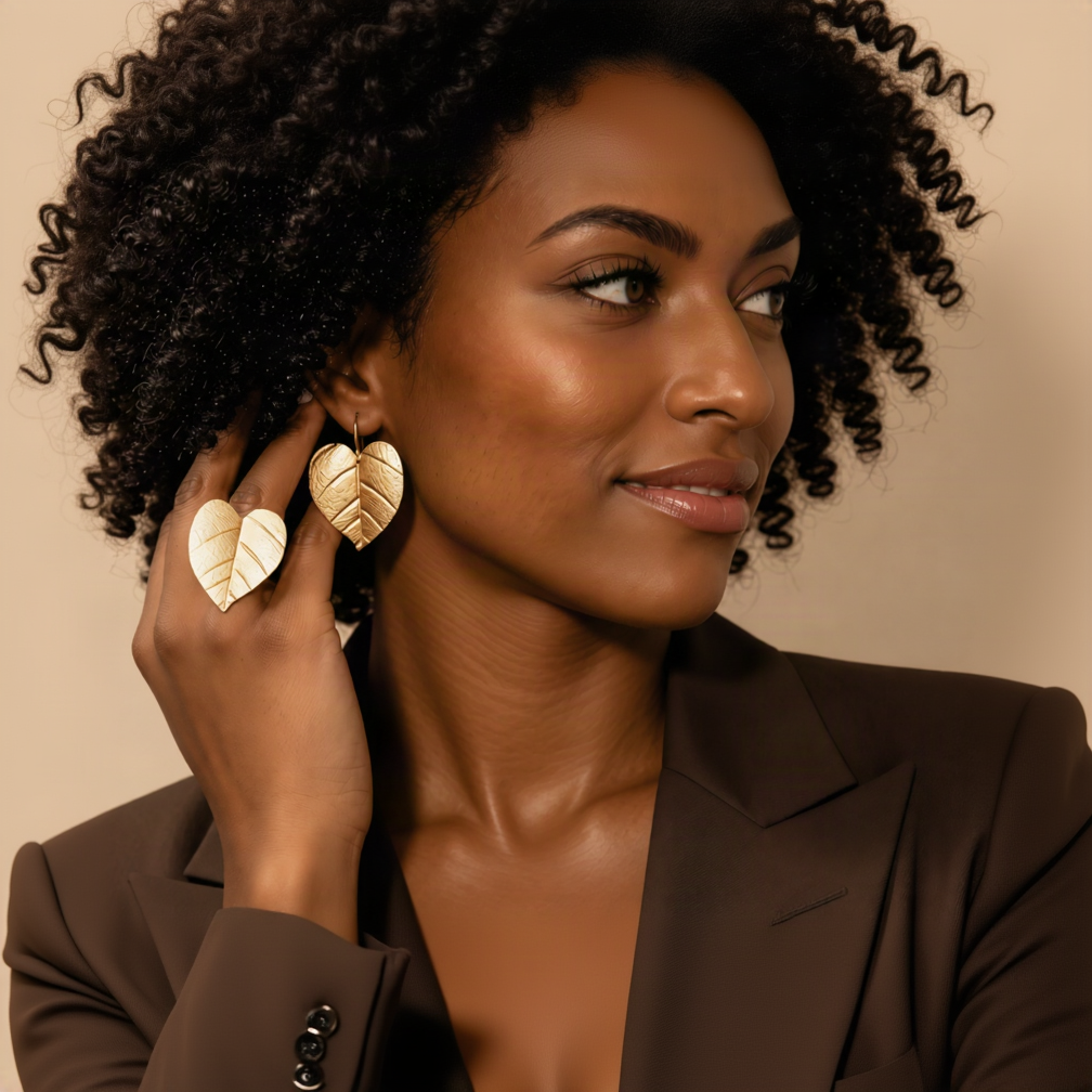 A close-up of a woman with curly black hair wearing gold leaf-shaped drop earrings and a matching gold ring on her finger, dressed in a dark brown blazer against a warm beige background.