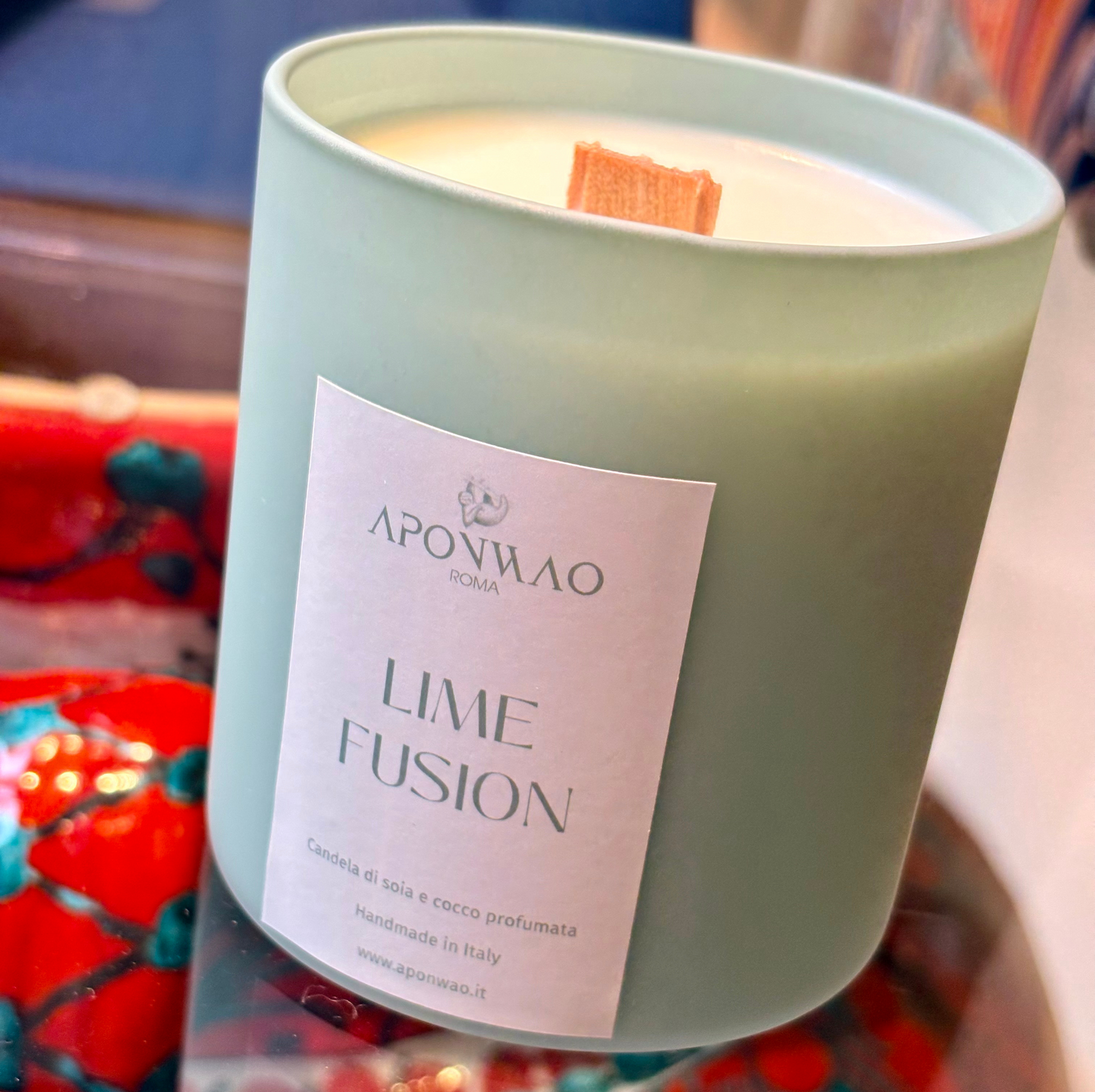 A close-up of a candle in a frosted sage green glass jar with a white paper label reading 'APONWAO' and 'LIME FUSION'. The candle has a light cream wax surface and a small orange wick.