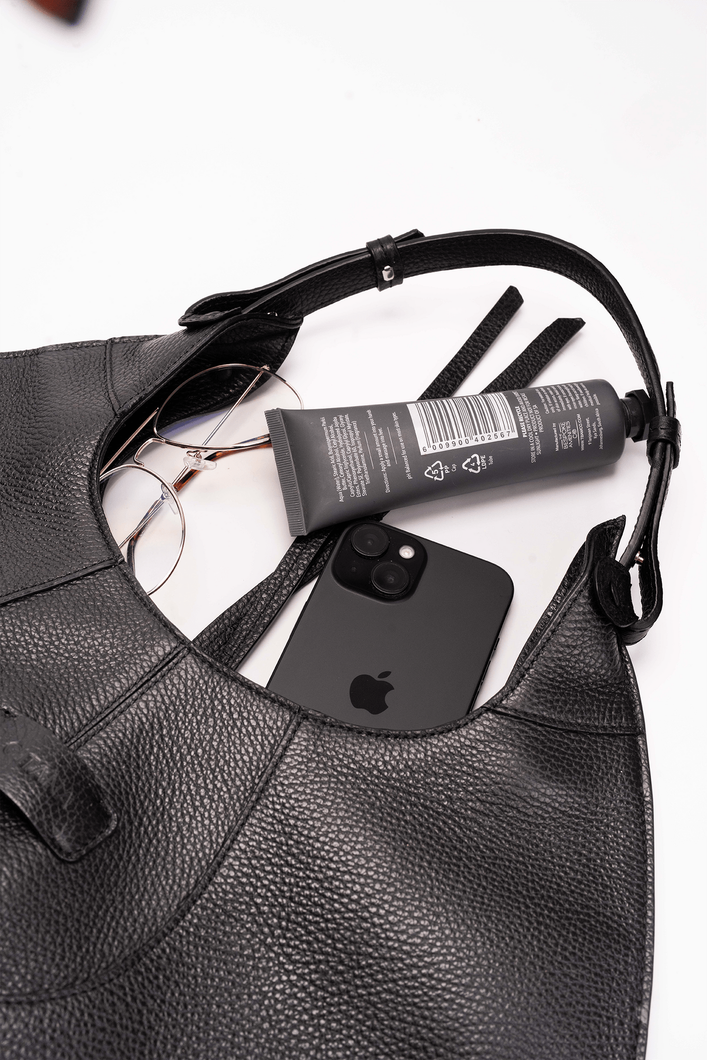 Black leather handbag with an iPhone, tube of cream, and sunglasses on a white background