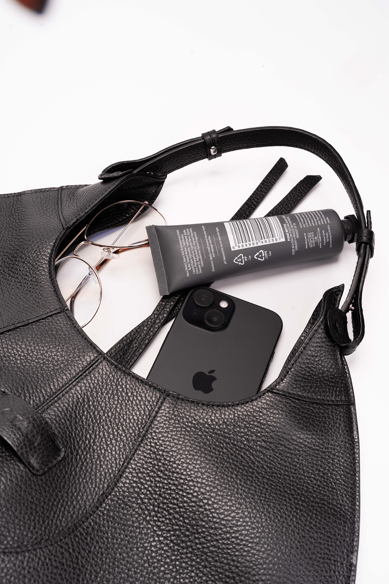 Black leather handbag with an iPhone, tube of cream, and sunglasses on a white background