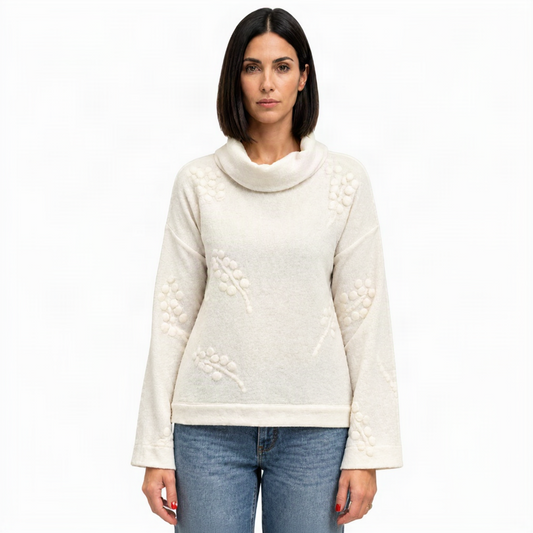 A woman modeling a cream-colored knit sweater with a cowl neck and raised floral embroidery, paired with blue jeans, against a plain white background.