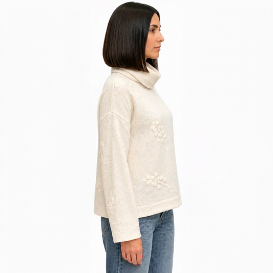 A woman shown in profile wearing a cream-colored knit sweater with floral embroidery and a high folded collar, paired with blue jeans, against a plain white background.