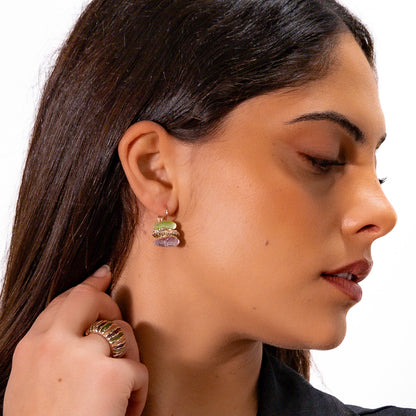 A close-up side profile of a woman wearing a small multicolor stud earring with stacked pastel green, pink, and purple elements, along with a textured gold ring on her finger, against a plain white background.