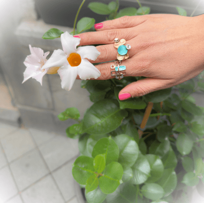 A close-up of a hand with pink nail polish wearing two rings, posed against a background of green leaves and white flowers. The rings feature gold-toned metal with turquoise and light blue stones.