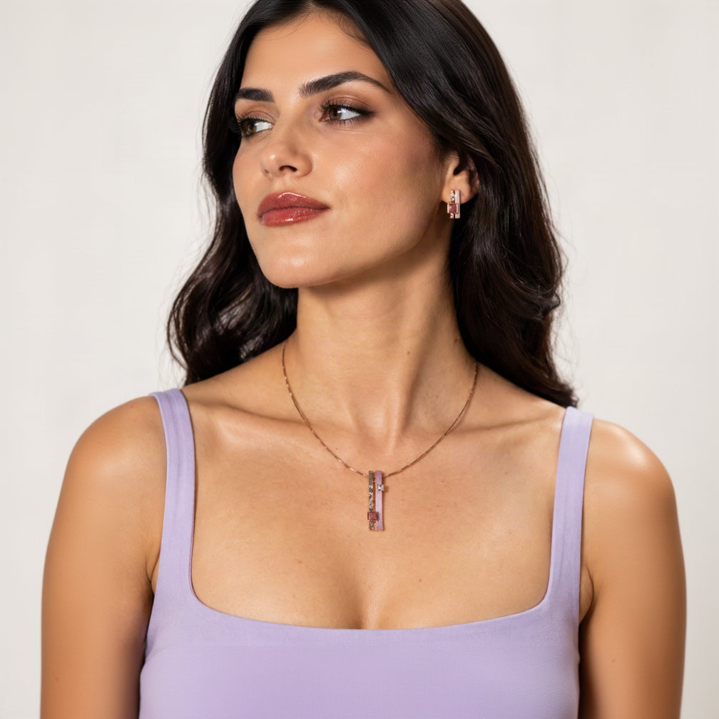 A close-up of a woman wearing A pendant of two slim bars, one hammered rose gold, one hand-painted in soft lilac, hanging side by side from a brass cable necklace that's twisted on itself for extra shine and character and matching rose gold stud earring, paired with a lilac vest strap top against a white background.