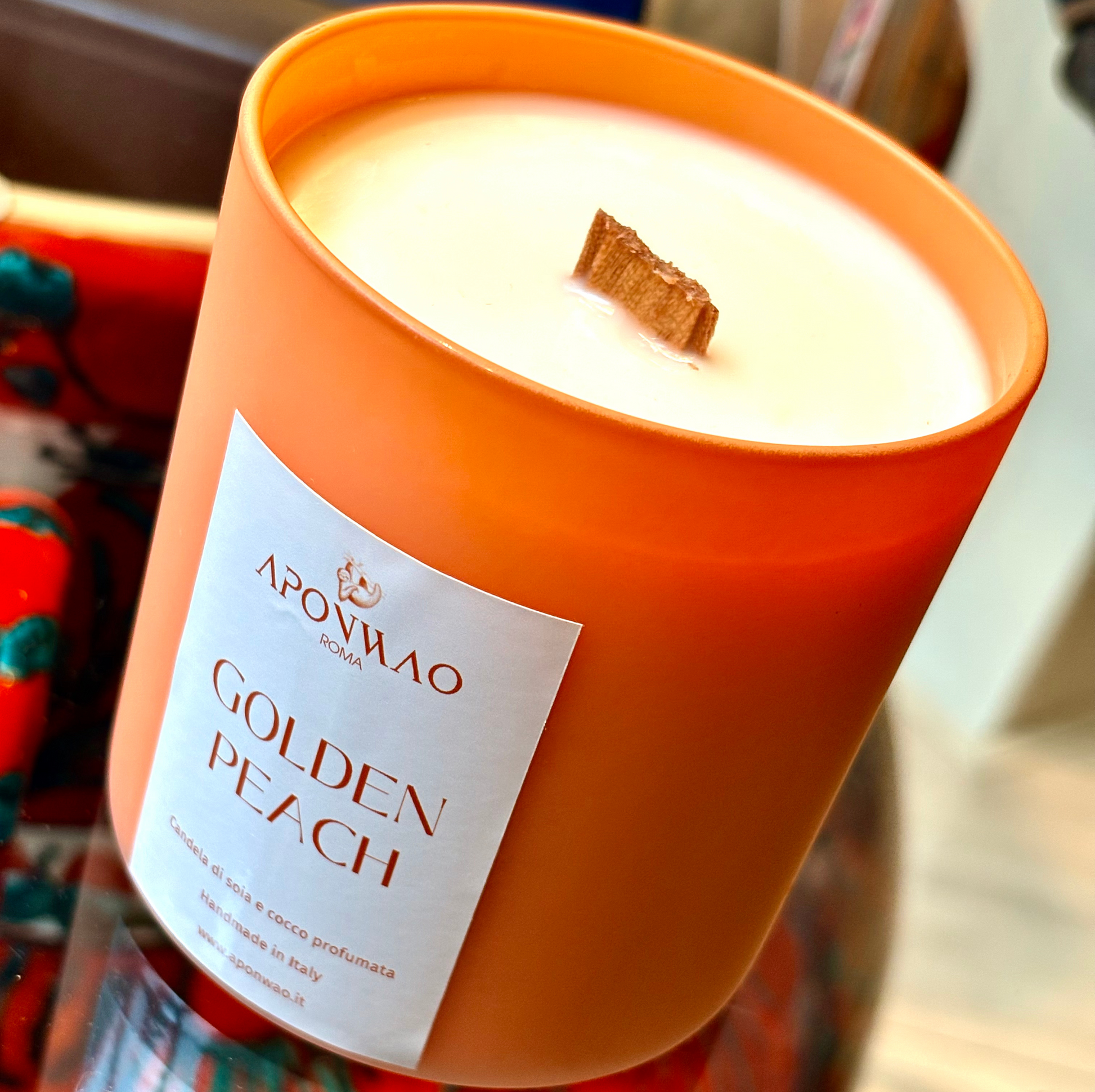 An orange glass candle jar with white wax and a wooden wick, featuring a white label that reads 'APONWAO GOLDEN PEACH'.