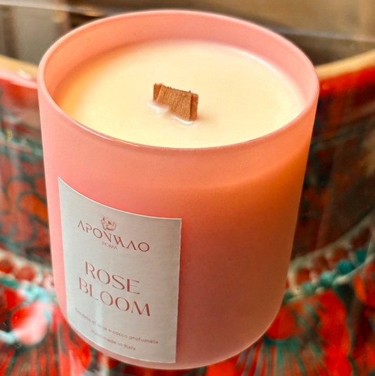 A pink glass jar candle with a white label reading 'APONWAO ROSE BLOOM' and a small wooden wick, set against a colourful patterned background.