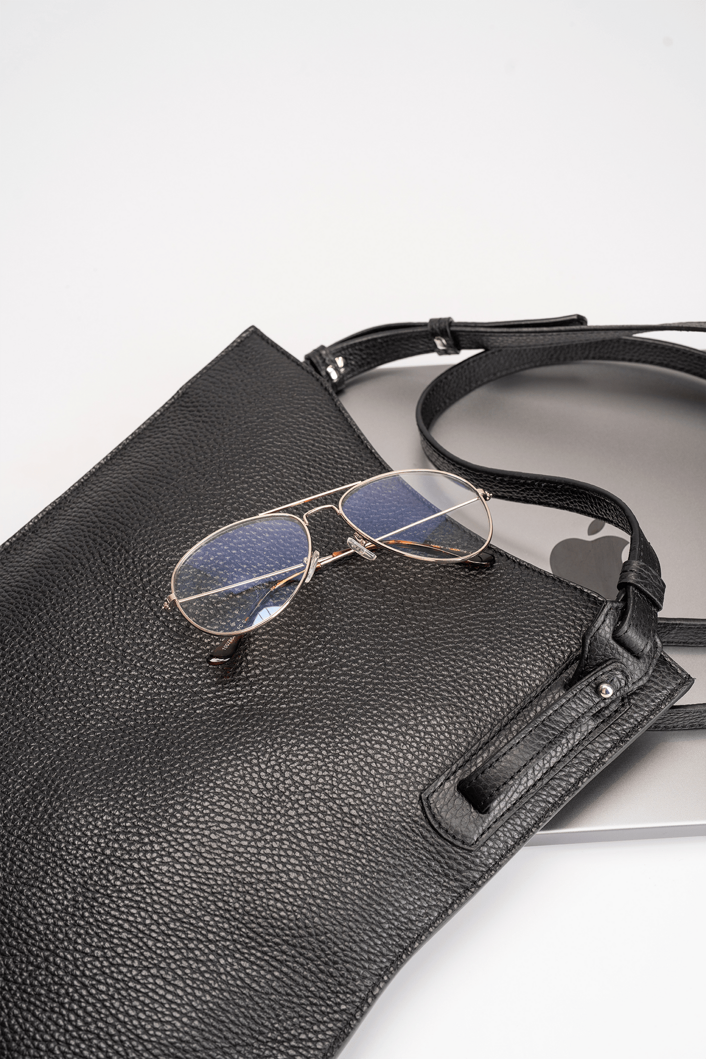 Black calf leather crossbody handbag displayed with MacBook and glasses on a white table to show bag capacity and lifestyle use.