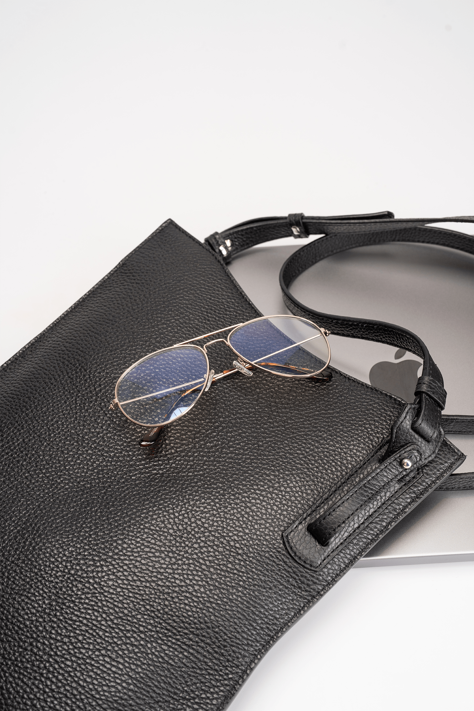 Black calf leather crossbody handbag displayed with MacBook and glasses on a white table to show bag capacity and lifestyle use.