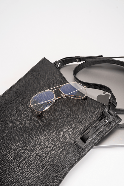 Black calf leather crossbody handbag displayed with MacBook and glasses on a white table to show bag capacity and lifestyle use.
